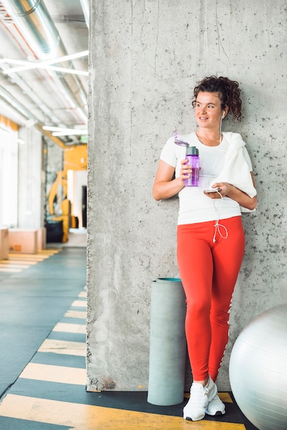 Happy woman with water bottle and smartphone leaning on wall in fitness club