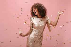 Free photo happy woman with wavy dark hair style in shiny trendy clothes rejoicing holding glass with champagne and posing with confetti on pink backdrop