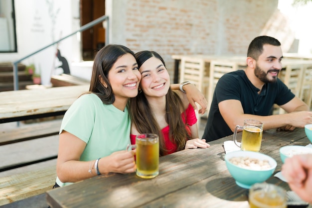 幸せな女性の親友は笑顔でアイコンタクトをしながら抱き締めます。ぶらぶらして屋外でビールを飲む友人のグループ