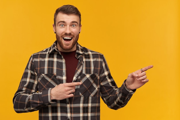 Happy wondered young man in plaid shirt with beard and opened mouth looks amazed and pointing away at empty space by two fingers over yellow wall