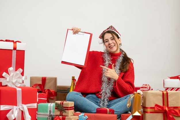 happy xmas girl with santa hat holding papers sitting around presents on white