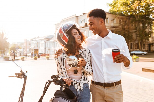 路上のモダンなバイクの近くに立って、お互いに見ながらコーヒーを飲みながら幸せな若いアフリカカップル