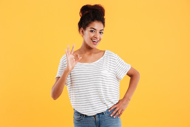 Happy young african lady in white pullover showing ok gesture and smiling isolated over yellow