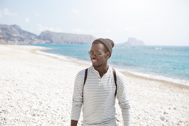 Happy young Afro American traveler in stylish hat and sunglasses having nice walk along seashore, enjoying sunny weather and beautiful views. Attractive young black man posing in sea scenery