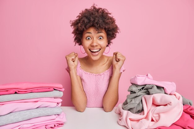 Happy young Afro American woman folds laundry at home poses at table with pile of folded and folded clothes clenches fists from joy as almost finished domestic work isolated over pink wall