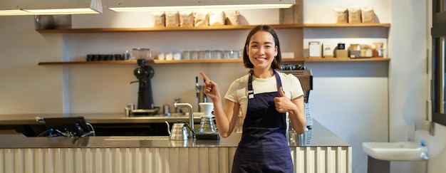 Foto gratuita felice giovane barista asiatico lavoratrice del personale in grembiule che mostra i pollici in su e indica lo spazio della copia