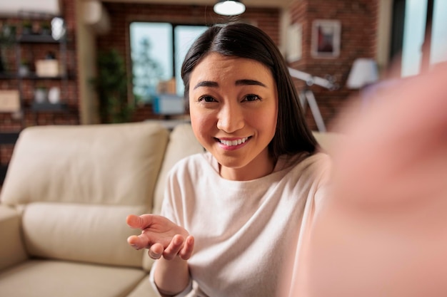 Felice giovane donna filippina cinese asiatica seduta sul divano a casa tenendo il telefono sullo schermo agitando la mano videochiamando amico a distanza online nell'app di chat mobile utilizzando l'applicazione di videochat per smartphone