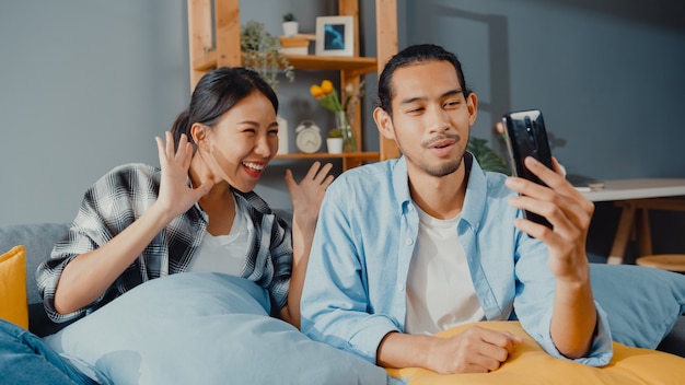 Happy young asian couple man and woman sit couch use smartphone facetime video call with friends and family