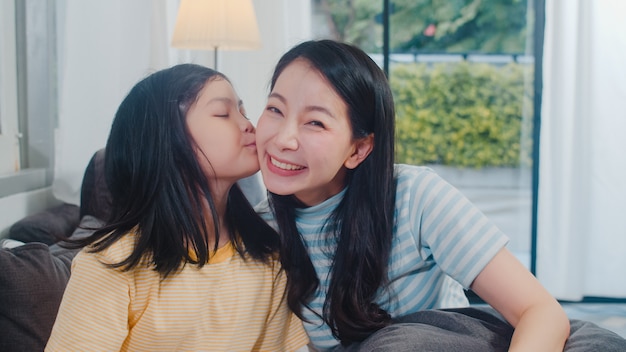 Happy young Asian family mom and kid play together on couch at home. Child daughter kiss her mom enjoying happy relax spending time together in modern living room in evening.