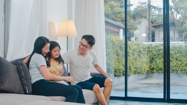 Happy young Asian family play together on couch at home. Chinese mother father and child daughter enjoying happy relax spending time together in modern living room in evening.