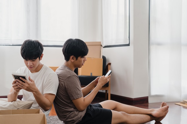 Happy young Asian gay couple relocation removals settle in new home. Asia lover guy lgbtq+ open cardboard box or parcel unpacking in living room on moving day. Real estate dwelling, loan and mortgage.
