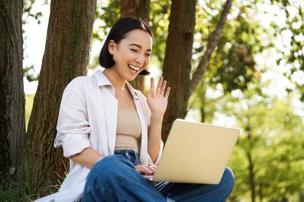 행복한 젊은 아시아 소녀는 나무 근처 공원에 앉아 야외에서 원격으로 작업하는 노트북을 보고 있습니다.