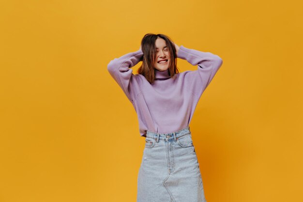Free photo happy young asian girl smiles sincerely and ruffles hair on isolated pretty woman in denim skirt and purple sweater poses on great mood on orange background