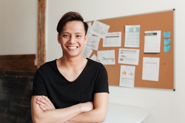 Happy young asian man standing indoors with arms crossed