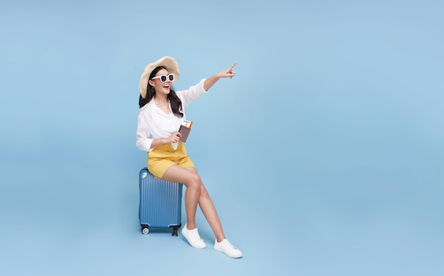 Happy young Asian tourist woman sitting on luggage holding passport and hand pointing to copy space