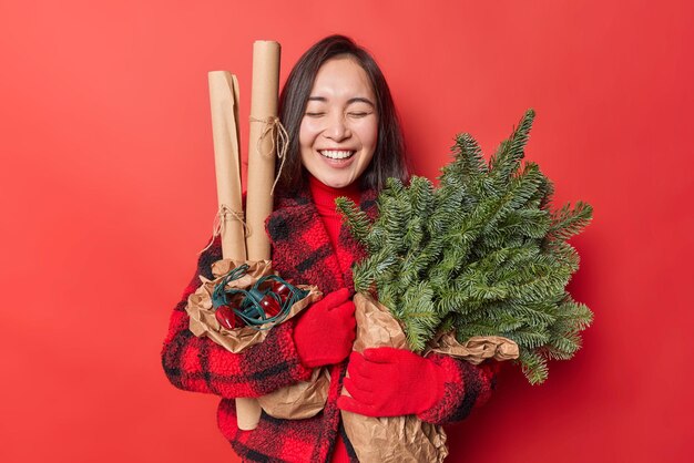 Happy young Asian woman laughs joyfully holds green spruce branches wrapped in paper retro garland returns from Christmas market prepares for holidays wears winter clothes isolated over red wall