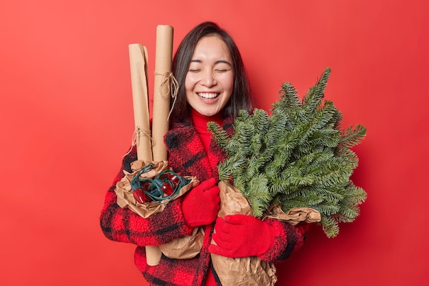 Happy young Asian woman laughs joyfully holds green spruce branches wrapped in paper retro garland returns from Christmas market prepares for holidays wears winter clothes isolated over red wall