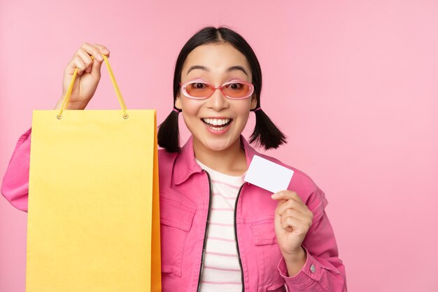 ピンクの背景の上に立っている店の店に行く販売中のバッグの購入を保持して買い物のためのクレジットカードを示す幸せな若いアジアの女性