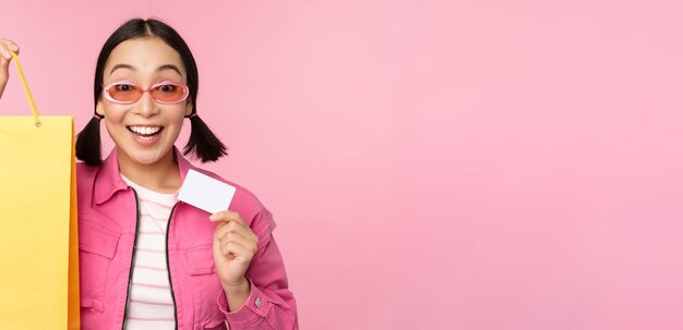 Happy young asian woman showing credit card for shopping holding bag buying on sale going to the shop store standing over pink background