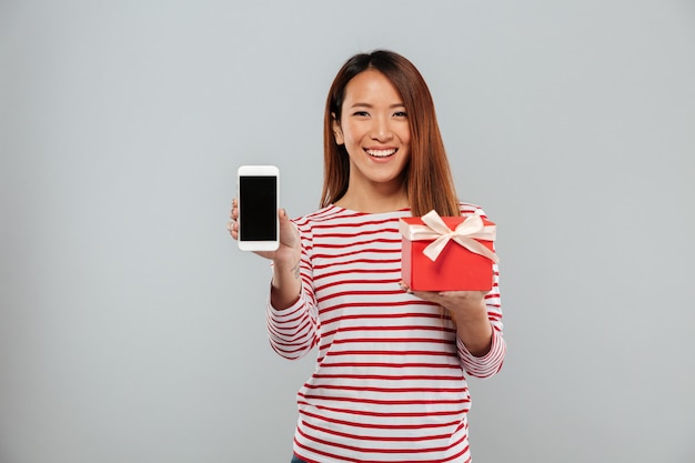 Happy young asian woman showing display of phone holding gift.