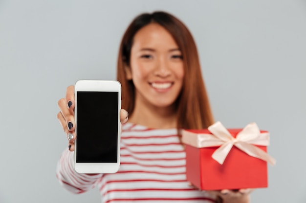 Happy young asian woman showing display of phone holding gift.