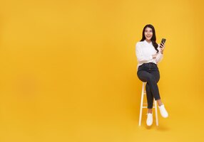 Счастливая молодая азиатка показывает мобильный телефон, сидя на белом стуле
