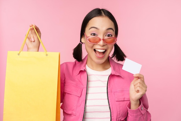 Happy young asian woman shows credit card and shopping bag store sale announcement buying smth in shop posing against pink background