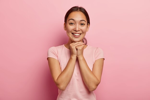 Happy young Asian woman with combed dark hair, keeps hands together under chin, glad to hear good news, has charming look, wears casual rosy t shirt, expresses good emotions, has piercing in ear