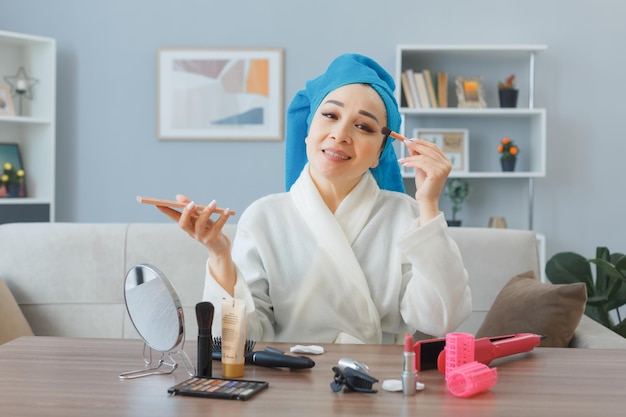 Felice giovane donna asiatica con un asciugamano in testa seduto al tavolo da toeletta a casa interni applicando ombretti facendo routine di trucco mattutino