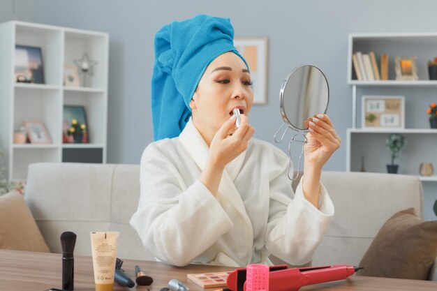 Happy young asian woman with towel on her head sitting at the dressing table at home interior looking at mirror applying lipstick doing morning makeup routine