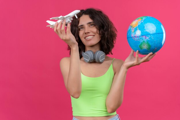 A happy young attractive woman with short hair in green crop top in headphones holding globe and looking at toy plane