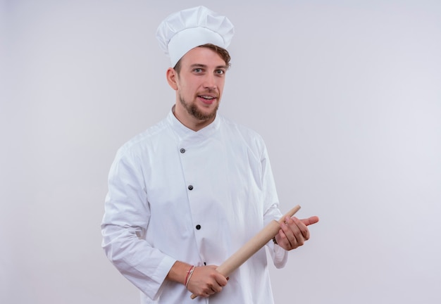 A happy young bearded chef man wearing white cooker uniform and hat holding rolling pin while looking on a white wall