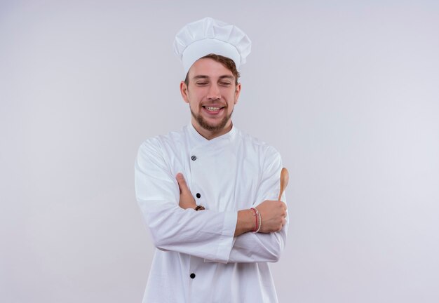 A happy young bearded chef man wearing white cooker uniform and hat holding wooden spoon with close eyes while standing on a white wall