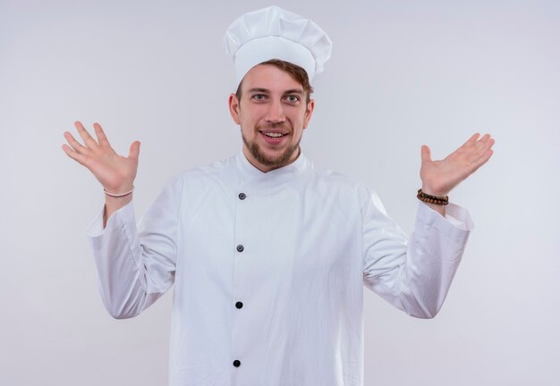 A happy young bearded chef man wearing white cooker uniform and hat opening his hands while looking on a white wall