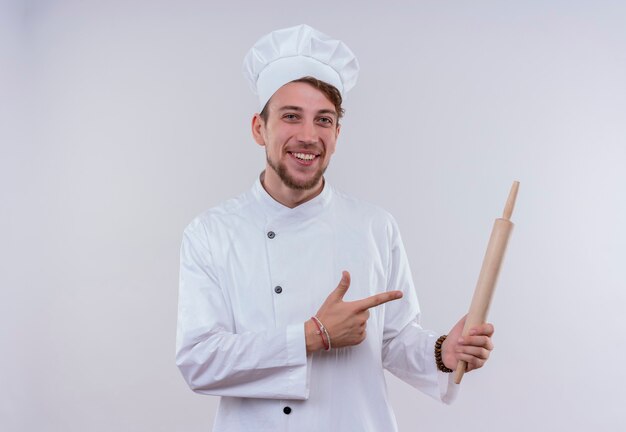 A happy young bearded chef man wearing white cooker uniform and hat pointing at rolling pin while looking on a white wall