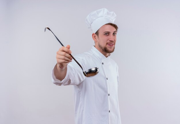 A happy young bearded chef man wearing white cooker uniform and hat showing ladle while looking on a white wall