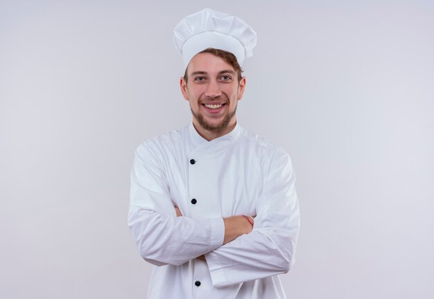 A happy young bearded chef man wearing white cooker uniform and hat smiling and holding hands folded while looking on a white wall