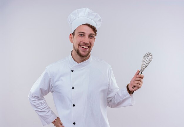 A happy young bearded chef man wearing white cooker uniform and hat smiling and holding mixer spoon while looking on a white wall