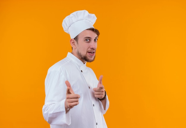 Un uomo felice giovane chef barbuto in uniforme bianca che punta con le dita indice su una parete arancione