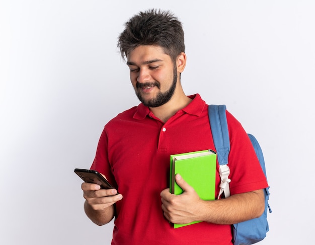 Happy young bearded student guy in red polo shirt with backpack holding notebooks holding smartphone looking at it smiling cheerfully standing over white wall