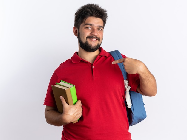 Happy young bearded student guy in red polo shirt with backpack holding notebooks pointing with index fingers at notebooks smiling cheerfully standing