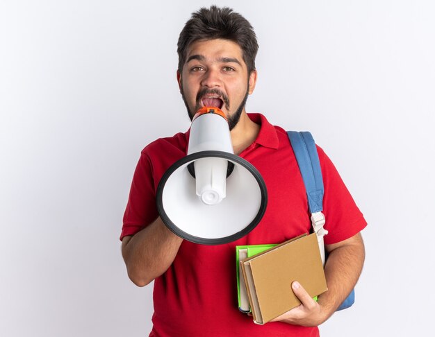 Happy young bearded student guy in red polo shirt with backpack holding notebooks shouting to megaphone standing