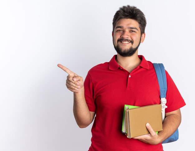 Happy young bearded student guy in red polo shirt with backpack holding notebooks  smiling cheerfully pointing with index finger to the side standing over white wall