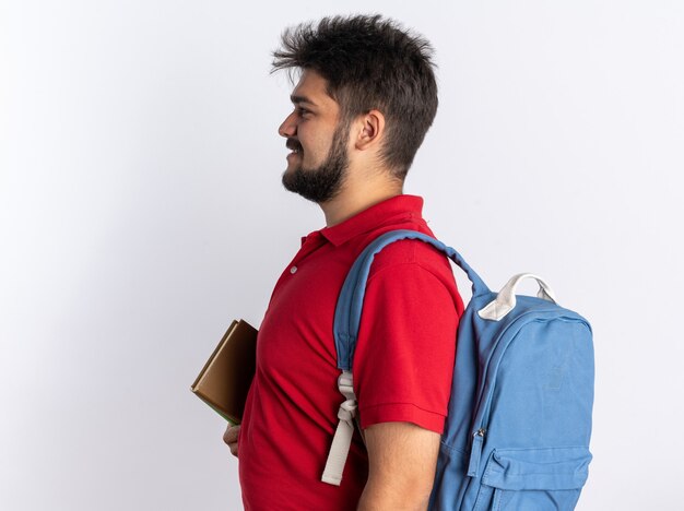 Happy young bearded student guy in red polo shirt with backpack holding notebooks standing sideways over white wall