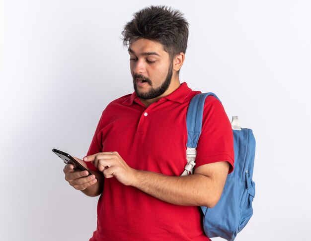 白い背景の上に立って笑顔のスマートフォンタイピングメッセージを保持しているバックパックと赤いポロシャツの幸せな若いひげを生やした学生男