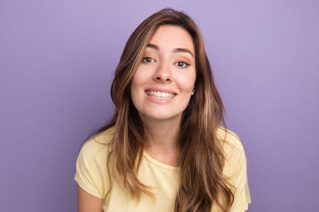 紫色の背景の上に広く立って笑顔のカメラを見てベージュのTシャツの幸せな若い美しい女性