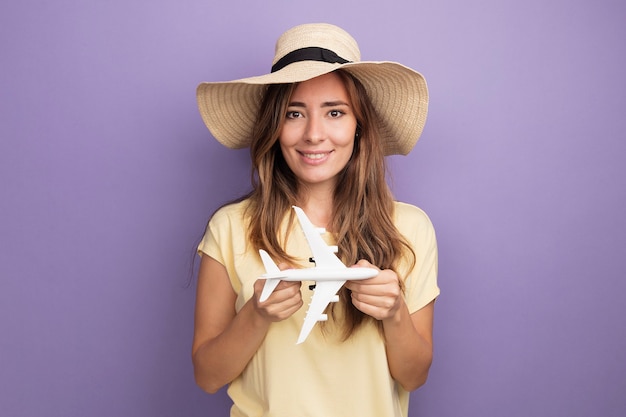 Happy young beautiful woman in beige t-shirt and summer hat holding toy airplane looking at camera with smile on face standing over purple background