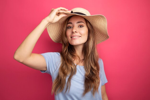 Happy young beautiful woman in blue t-shirt and summer hat looking at camera smiling cheerfully standing over pink background