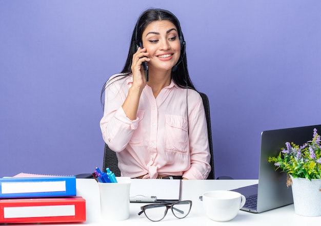 Счастливая молодая красивая женщина в повседневной одежде, носящей гарнитуру, уверенно улыбается во время разговора по мобильному телефону, сидя за столом с ноутбуком на синем фоне, работая в офисе