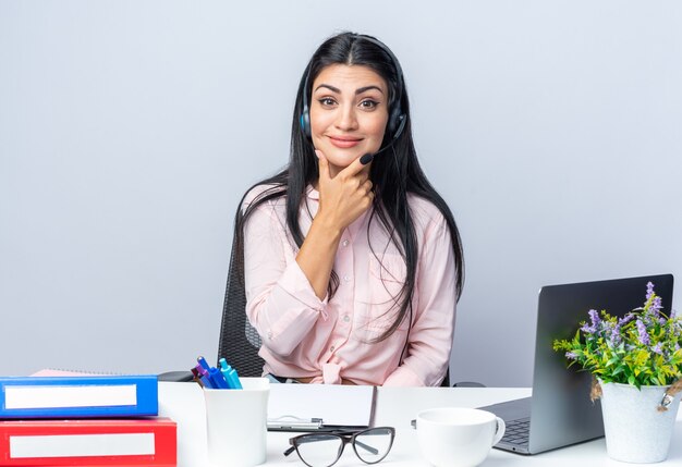 Happy young beautiful woman in casual clothes with headphones and microphone looking at front smiling confident sitting at the table with laptop over white wall working in office